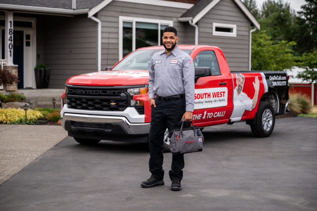 South West plumber holding tool bag while standing in front of service pickup truck parked in front of a gray home.