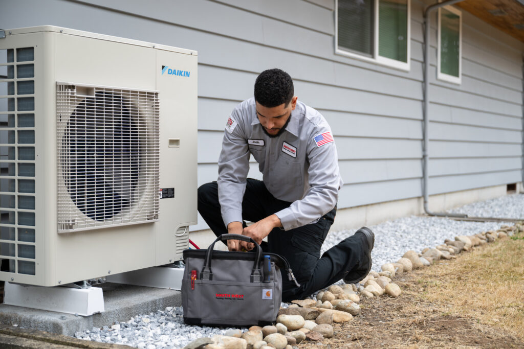 Technician with gray tool bag, repairing outdoor Daikin HVAC unit next to a home with light gray siding.