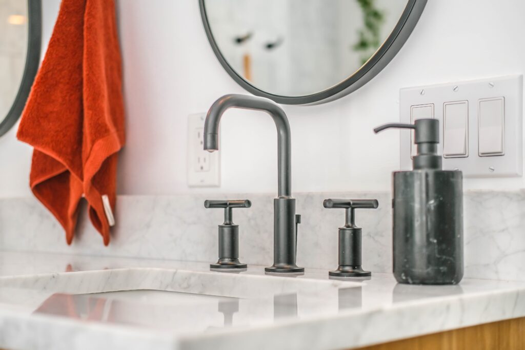 Gray bathroom faucet & sink in white stone countertop with orange hand towel handing on wall in background.