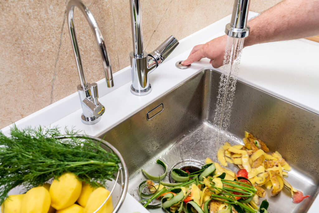 Person's hand turning on a garbage disposal in a modern kitchen stainless steel sink to remove vegetable peels.